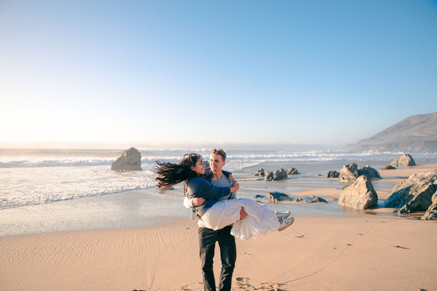 Man lifting woman on sunny beach with ocean waves and rocks in the background.