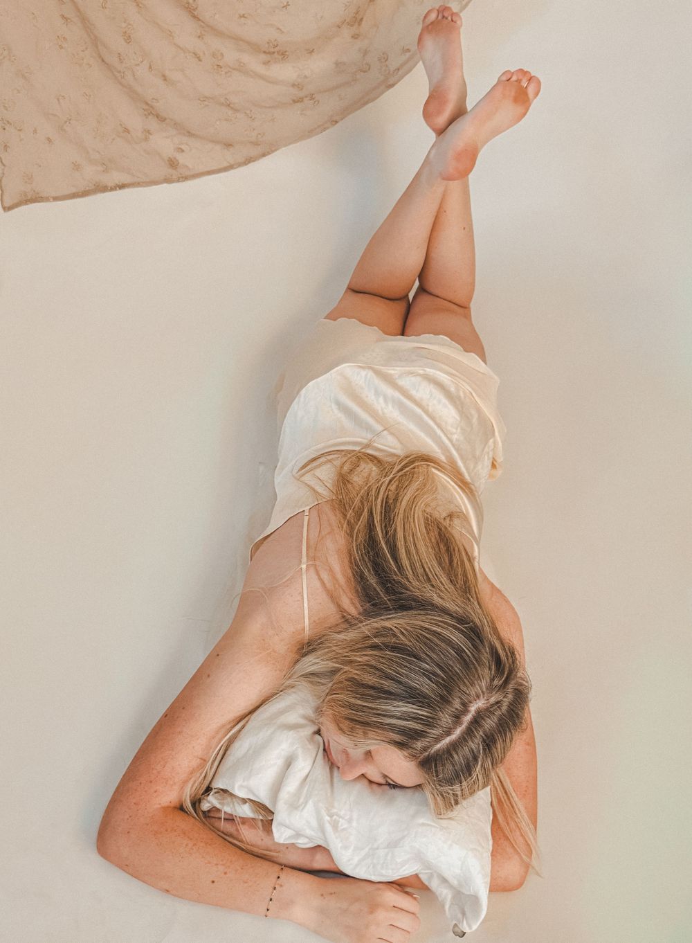 Overhead view of blonde woman in cream slip dress lying on white pillow with legs raised against neutral backdrop.