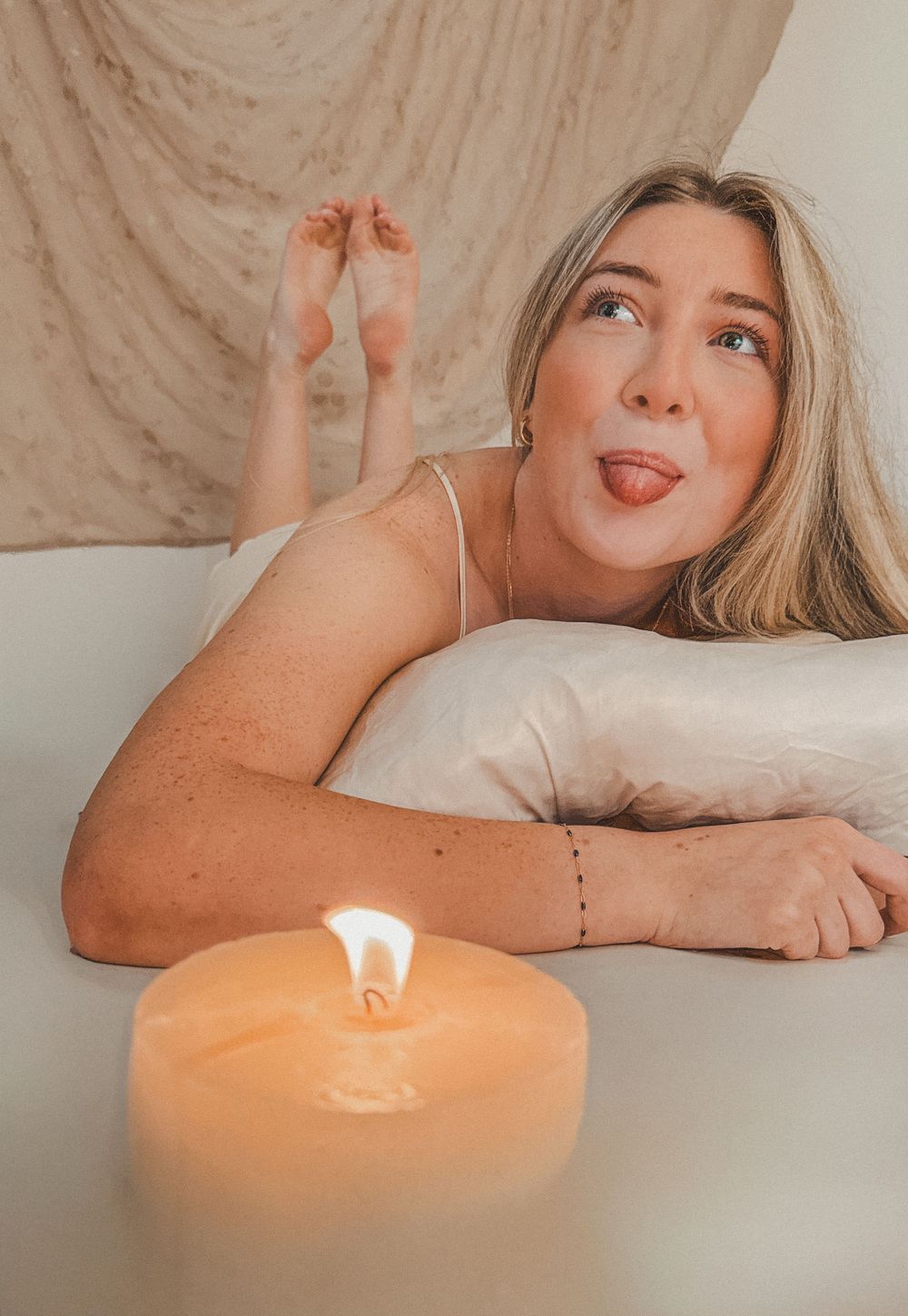 Playful blonde woman sticking out tongue while resting on pillow near glowing candle in cozy studio setting.