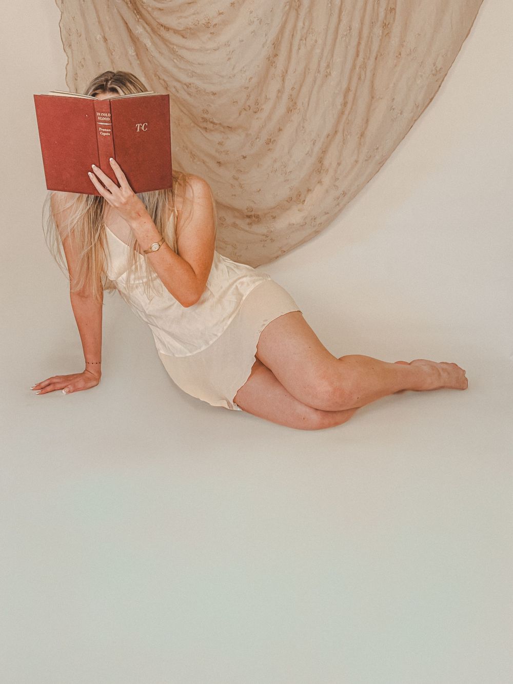Blonde woman in cream slip dress reading a red book while posing on floor with draped fabric backdrop.