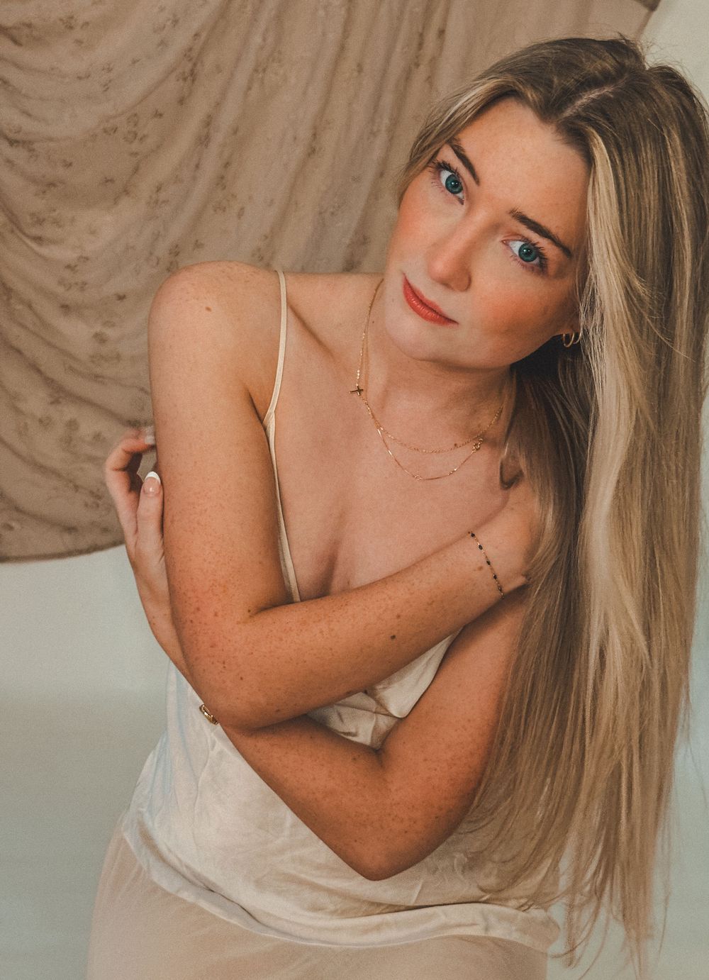 Blue-eyed blonde woman with freckles in cream slip dress crossing arms over chest in soft studio lighting.