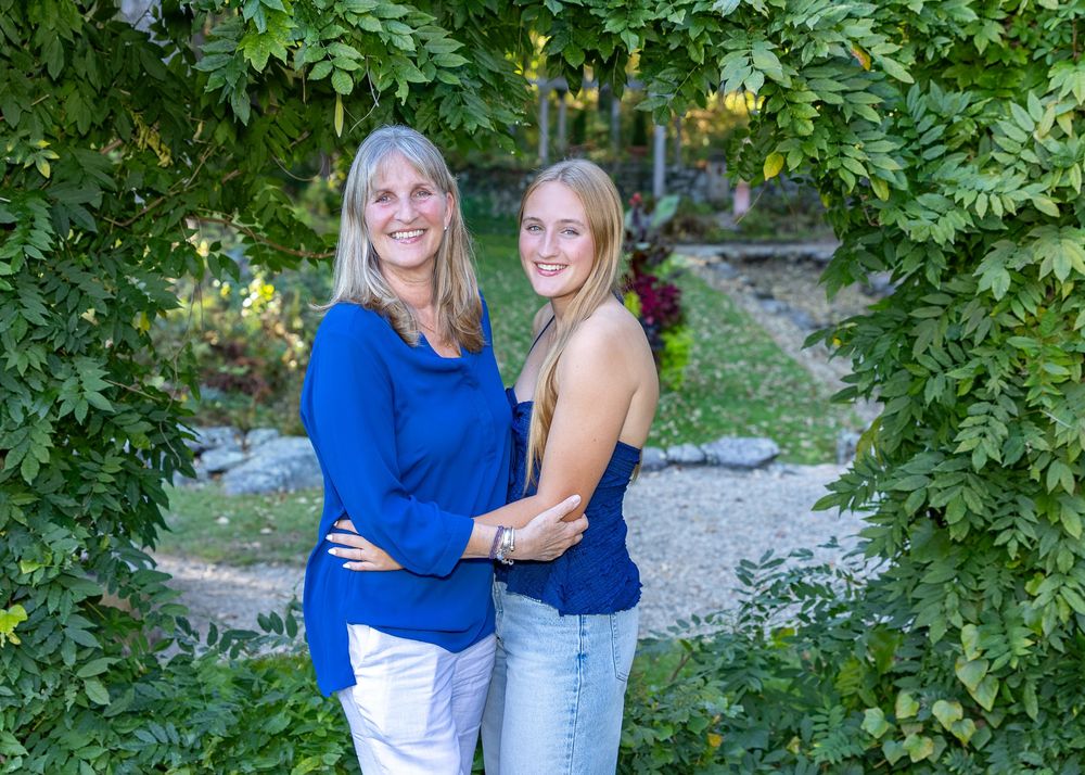 Mom & high school senior daughter in blue tops smile together in a lush garden archway, posing for an outdoor portrait in Lincoln, MA