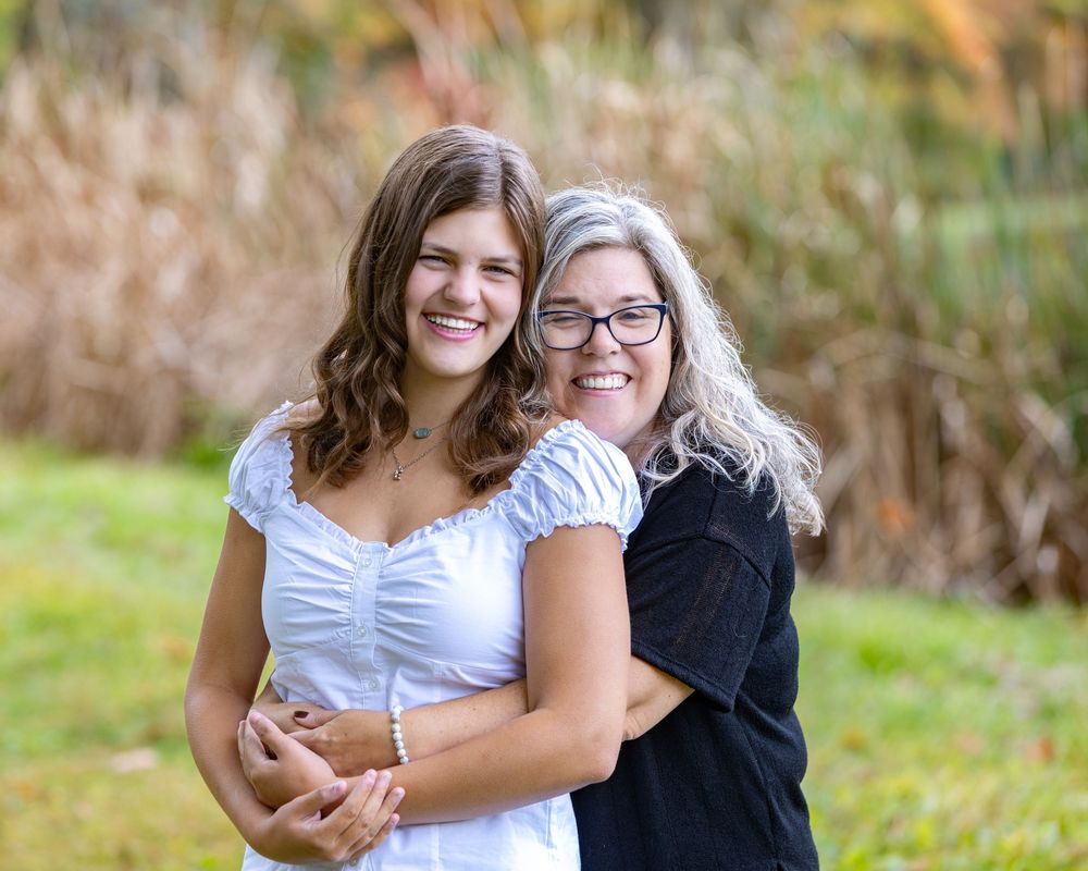 Mother and daughter embrace outdoors, one in white dress and one in black top, with autumn foliage background in Sudbury, MA.