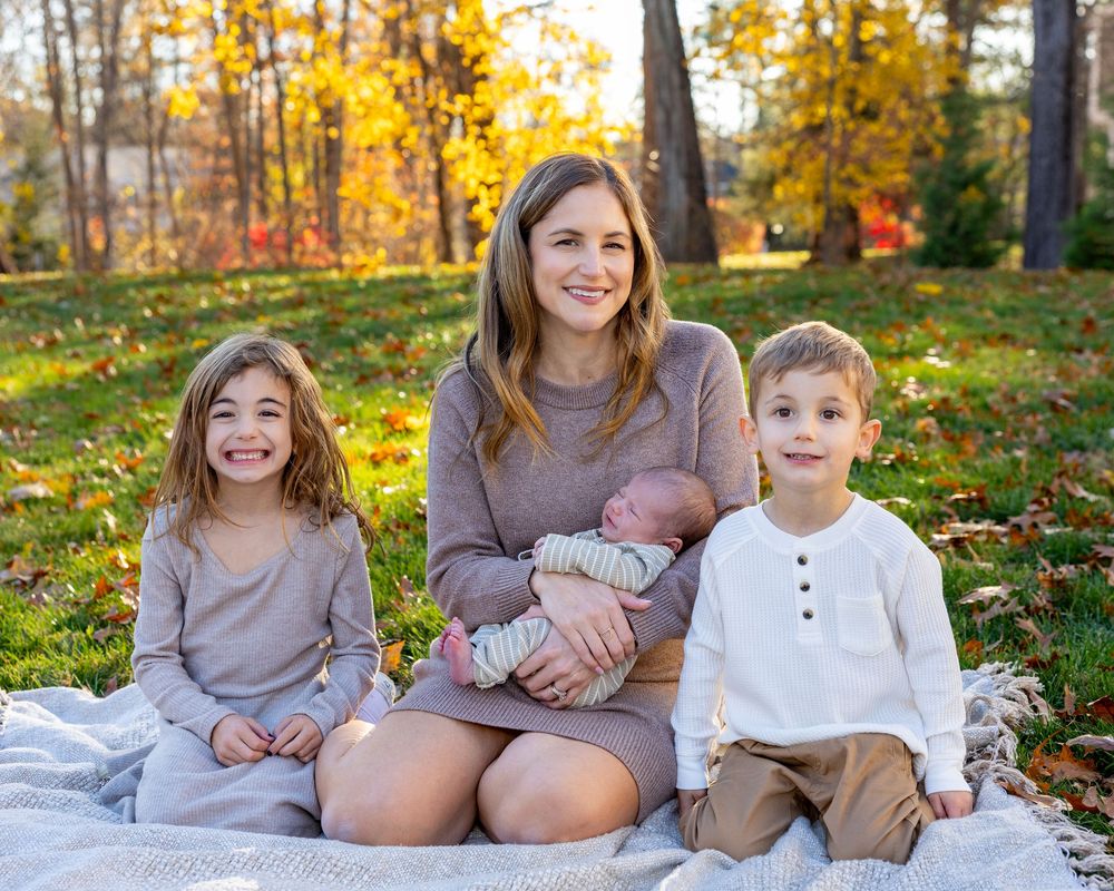 A mother sits outdoors on a blanket with three children, including a newborn, surrounded by autumn foliage in Stow, MA during family photos.