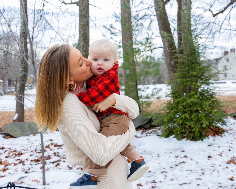 A mother kisses her baby boy in a red plaid outfit outdoors in a snowy winter setting during a family photo session in Concord, MA.