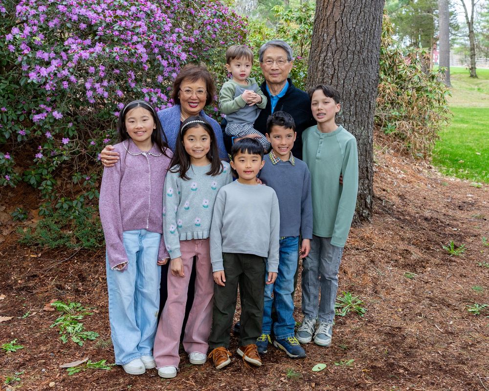 Grandparents and their grandchildren smile together outdoors near blooming purple azalea bushes and pine trees at multi-generational photos.
