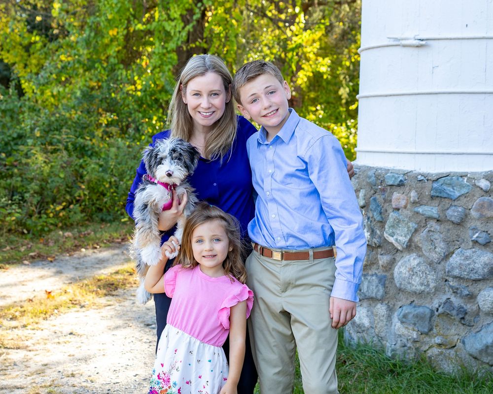 A smiling mother holds a small dog while posing outdoors with two children near a stone wall and trees during a family photo session.