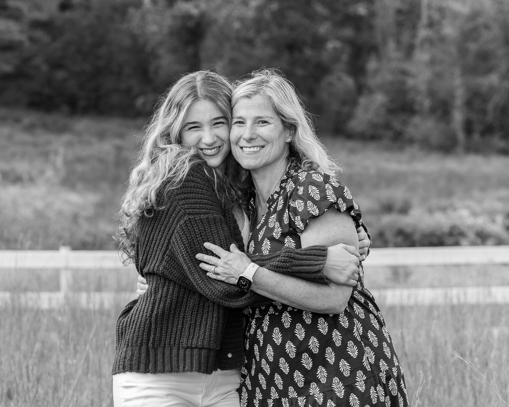 Mother & daughter pose smiling and hugging outdoors in a black and white portrait photo near a wooden fence in Sudbury, MA.