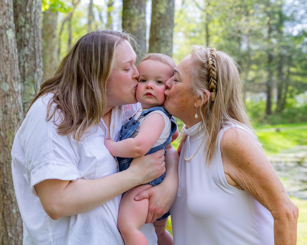 Mom & Grandmother kissing a happy baby outdoors in a lush green forest setting during a family photo session in Sudbury, MA.