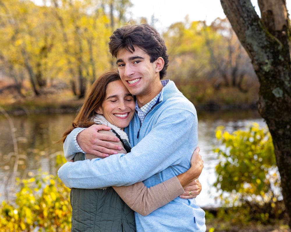 High school senior son & his mother pose near a river with colorful autumn foliage during a family photo session in Concord, MA.