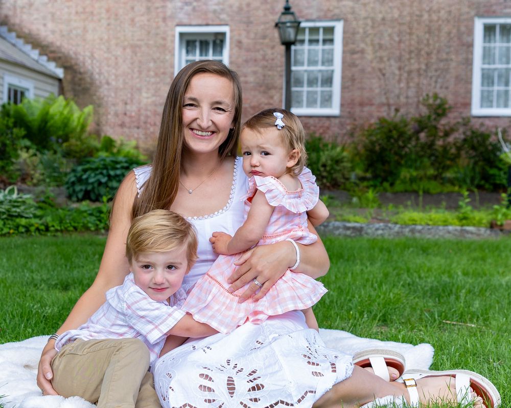 A smiling mother sits on a blanket outdoors holding a baby girl and a young boy in coordinating pink outfits during a family photo session.