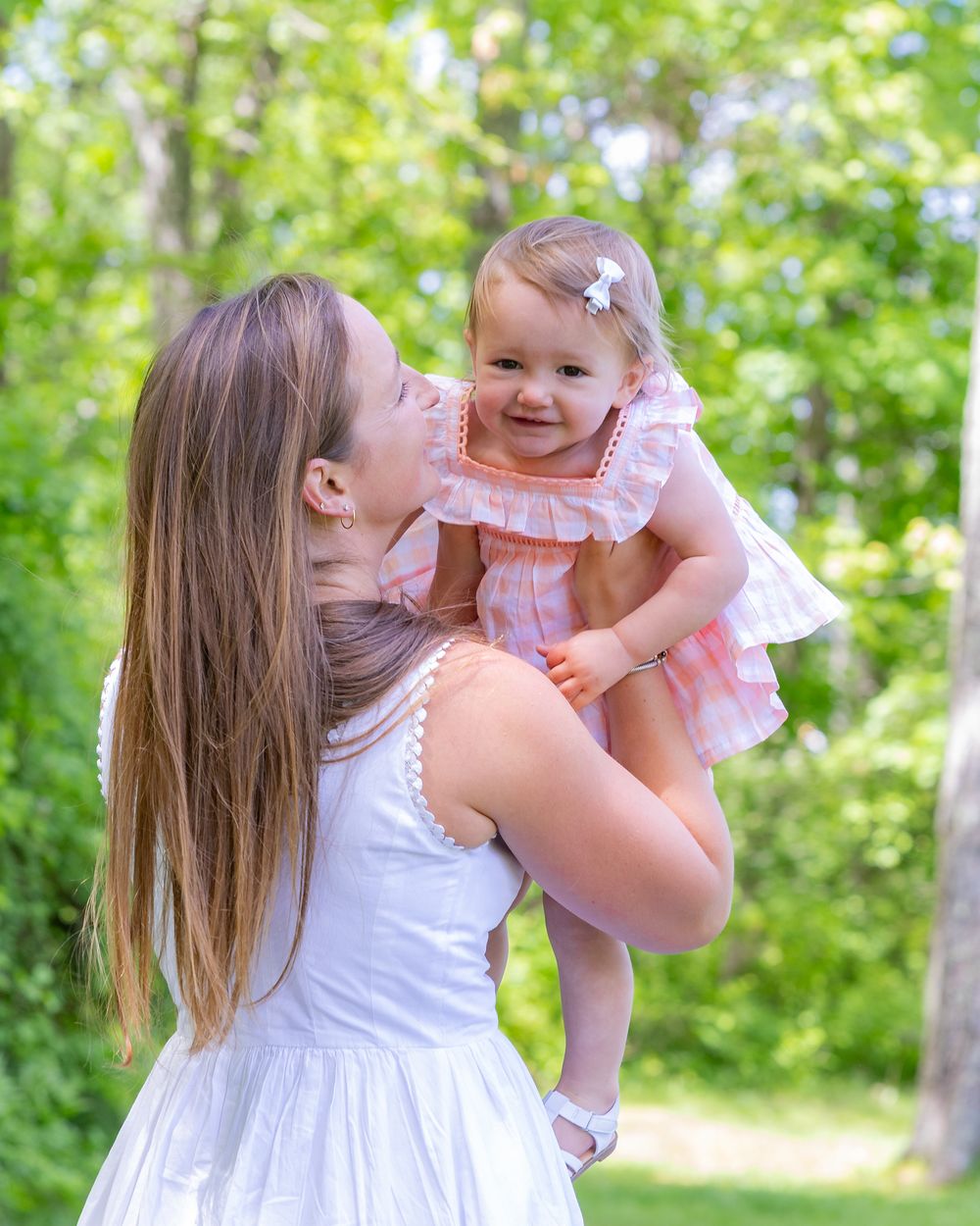 A young mother in a white dress holds her smiling baby girl in a pink dress among green trees during a family photo session in Sudbury, MA.