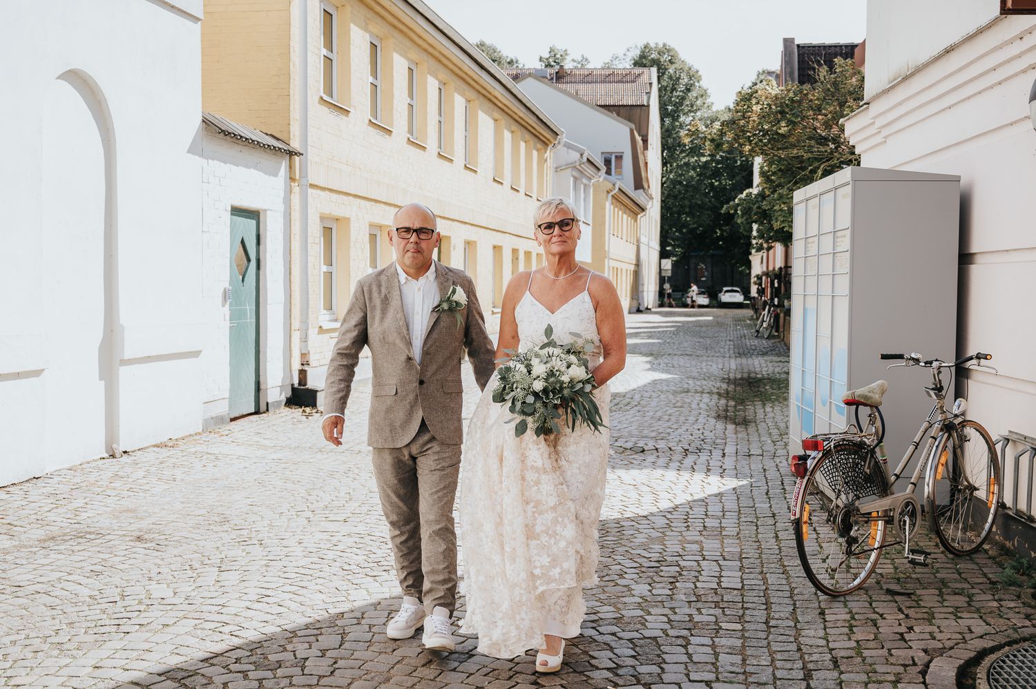 Bride and groom walking hand in hand along a sunny cobblestone plaza lined with trees on their wedding day.