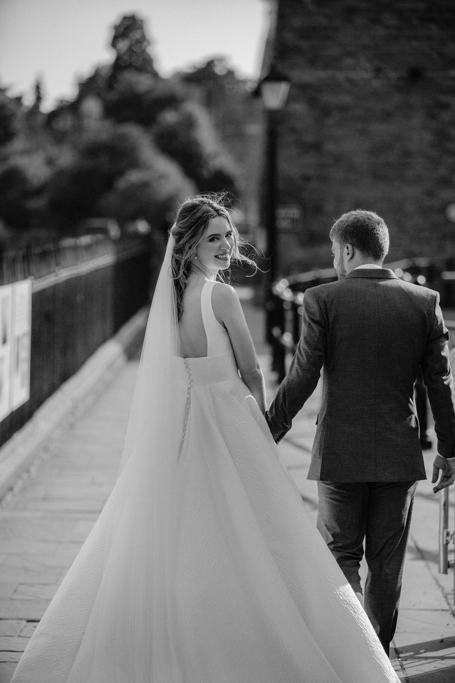 Black and white photo of bride and groom walking hand in hand on a cobblestone path, bride glancing back.