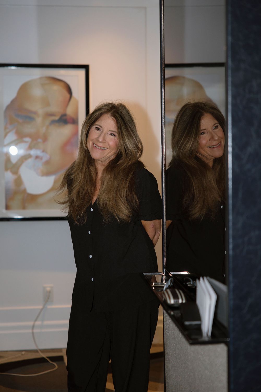 Reflection shot of a woman in black attire smiling against a gallery wall with artwork.