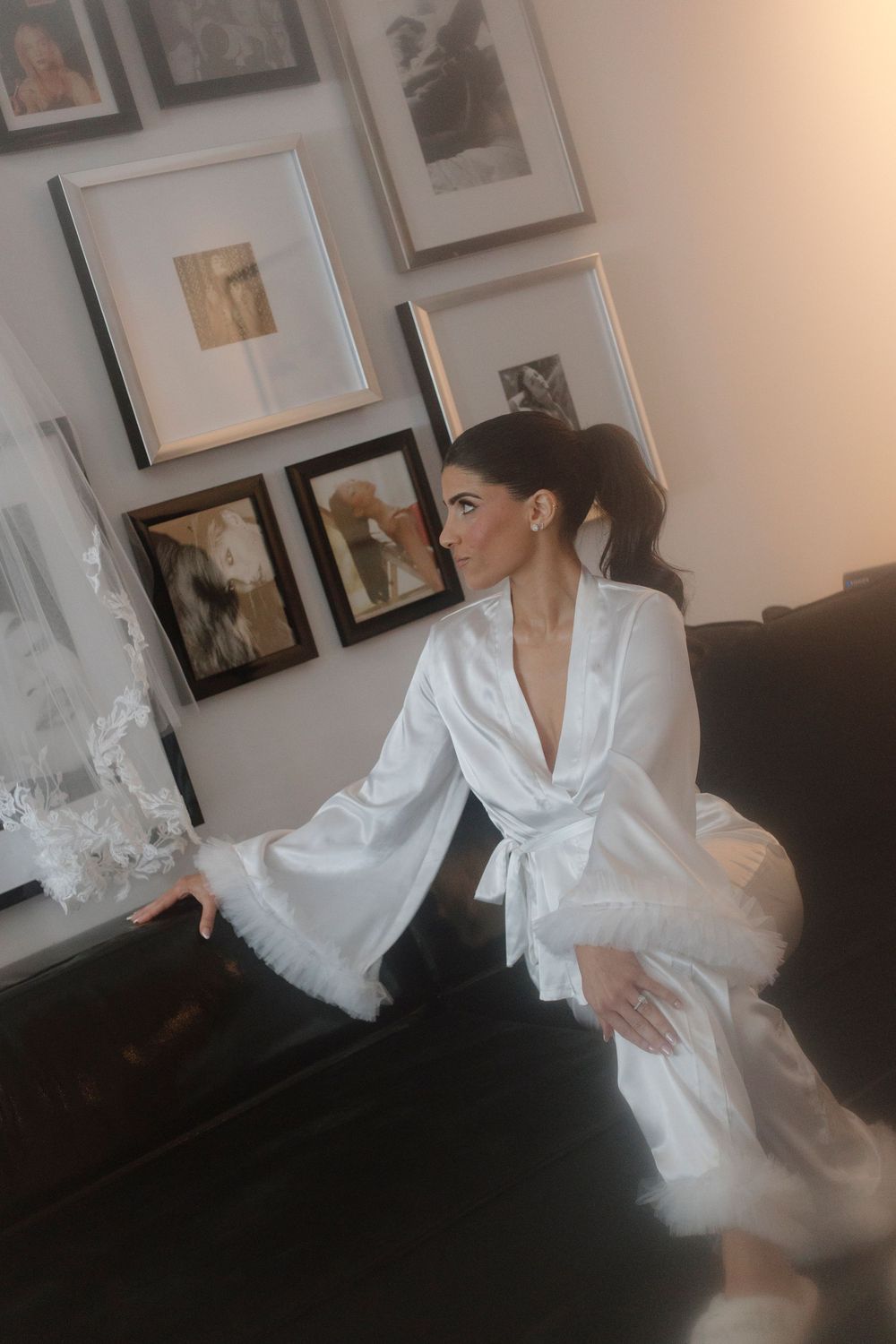 A person poses in an elegant white satin robe with dramatic bell sleeves against a backdrop of framed artwork.