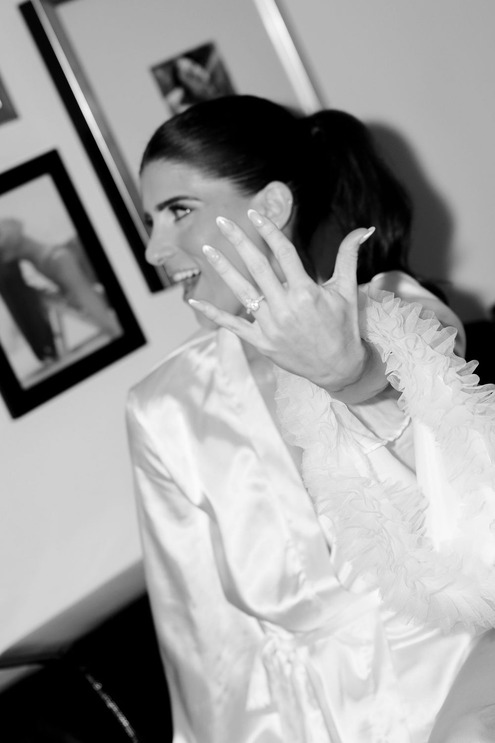 A bride in a white ruffled dress showing her ring with excitement.