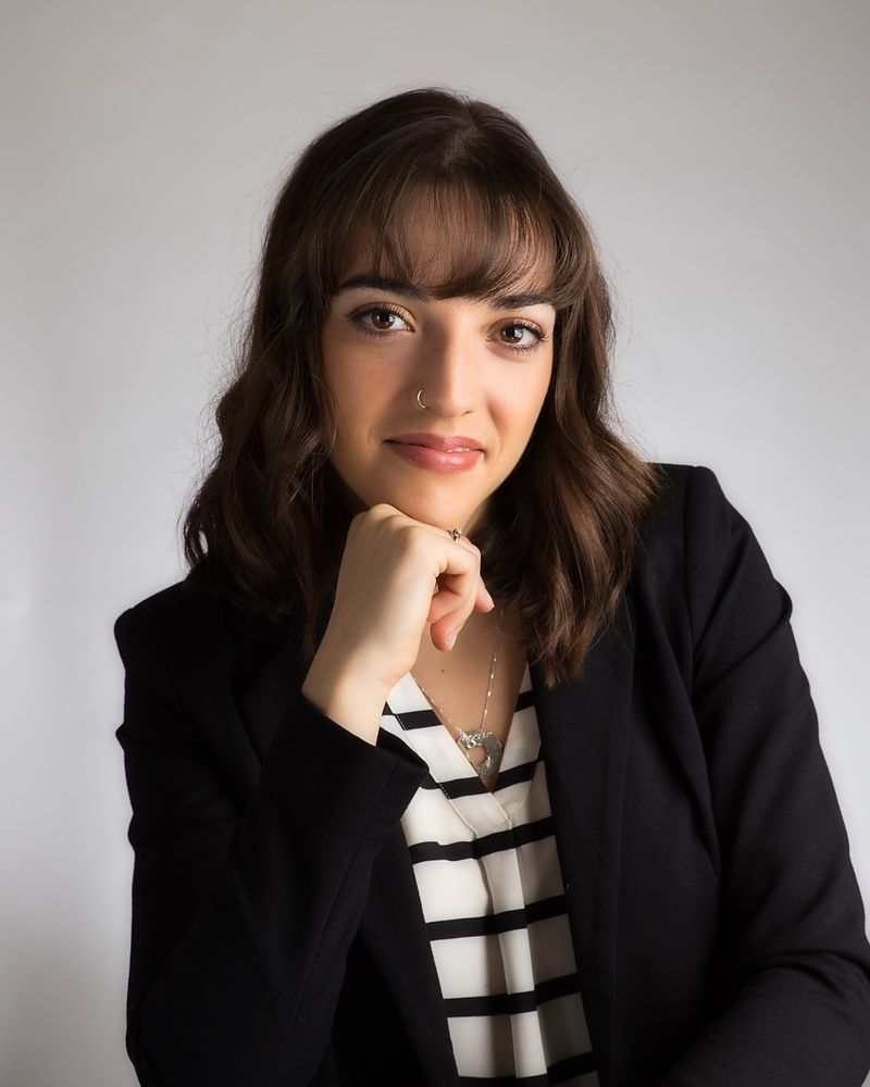 Professional portrait against neutral background showing a person in black blazer and striped shirt with natural lighting.