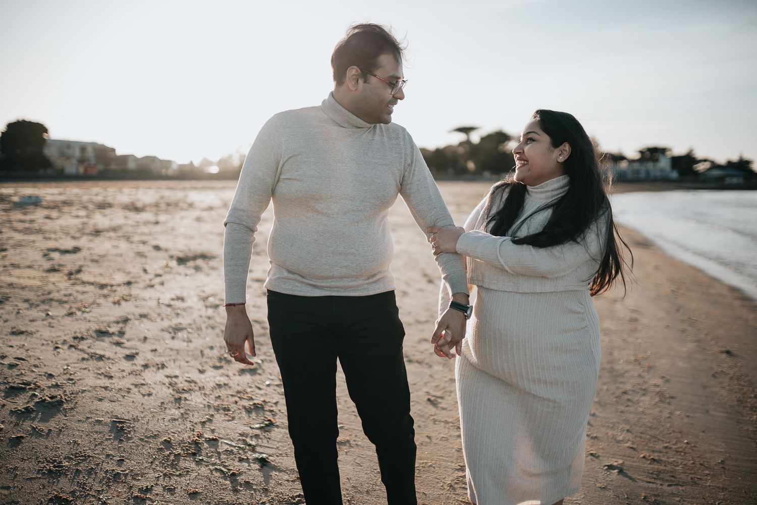 A couple in white sweaters walk together and smile on a sandy beach at sunset taken by Melbourne maternity photographer.