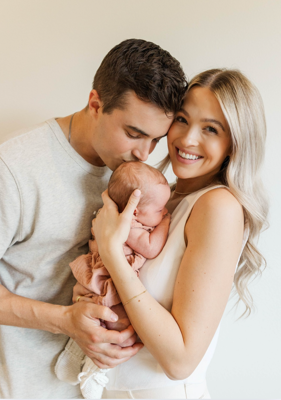 Loving couple sharing a tender moment with their newborn baby, father kissing infant while mother smiles warmly indoors.