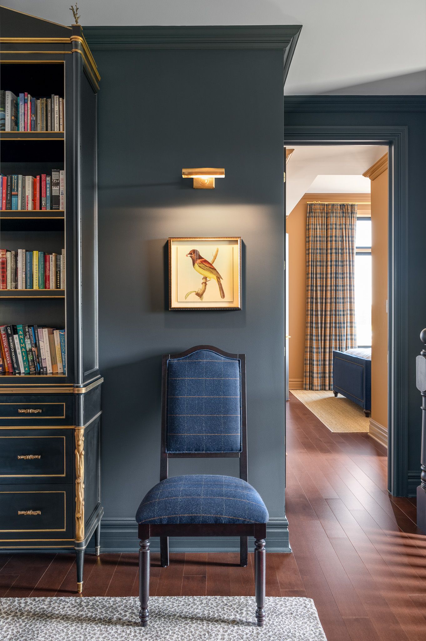 A stylish library corner with navy walls, built-in bookshelves, gold accents, and an upholstered blue chair beneath artwork.