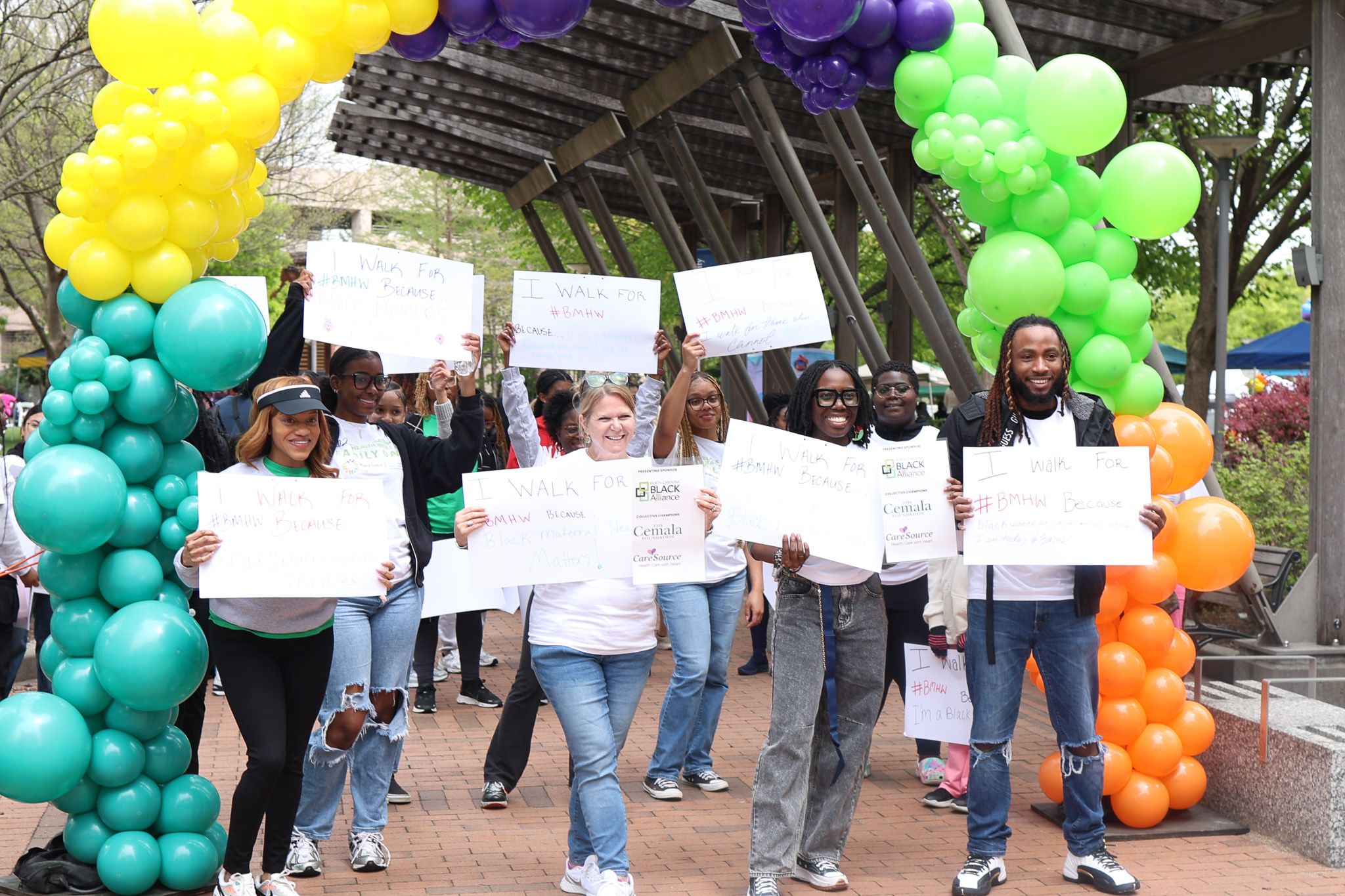 Rally participants march with signs under colorful balloon arch at outdoor advocacy event.