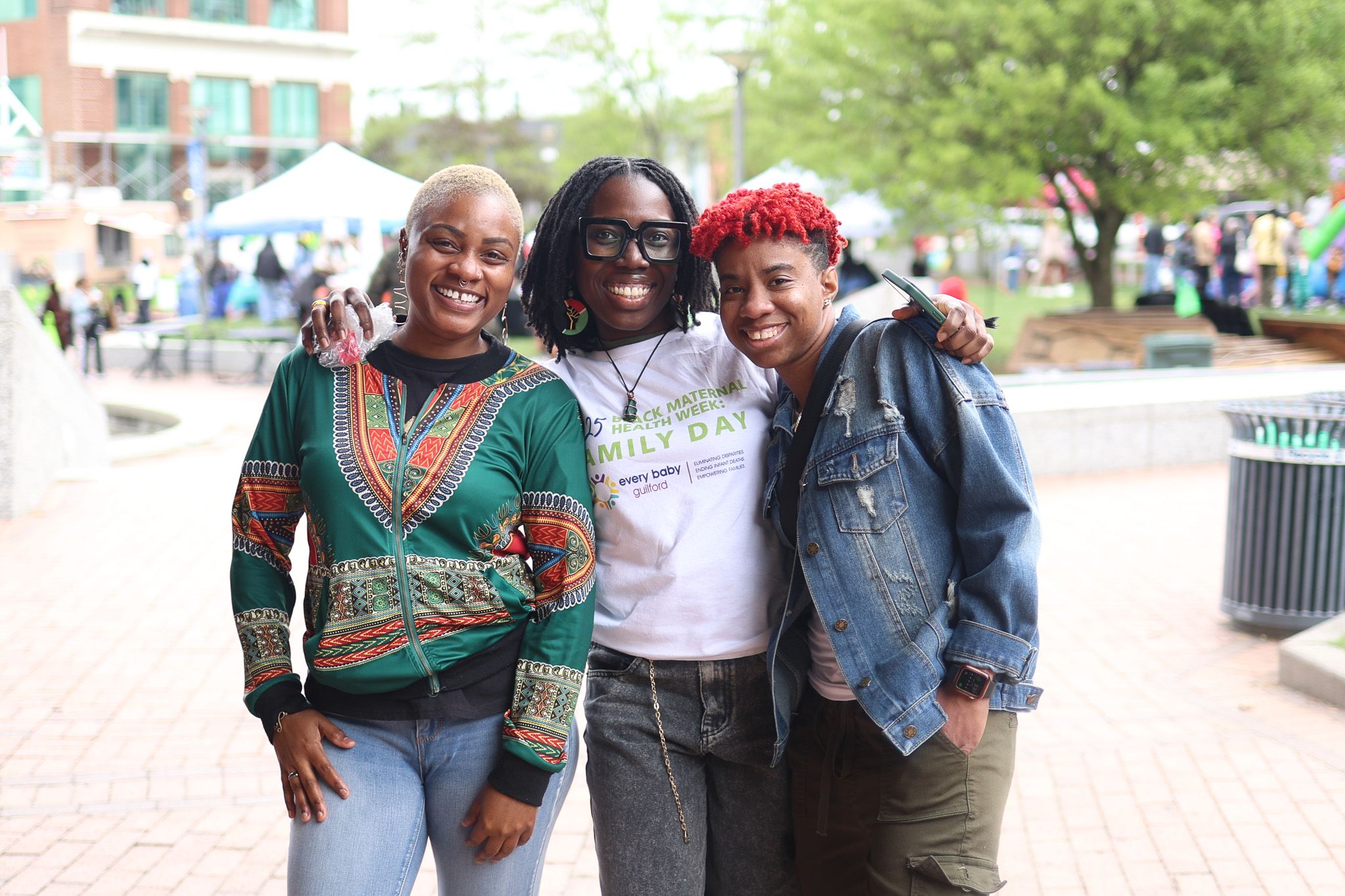 Three supporters smile together at outdoor community health event wearing colorful outfits.