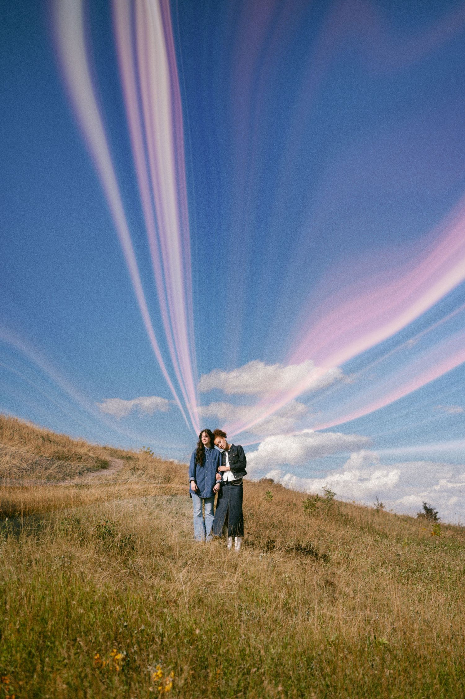 Two people hiking on a grassy hillside under dramatic pink and blue streaked clouds in the sky.