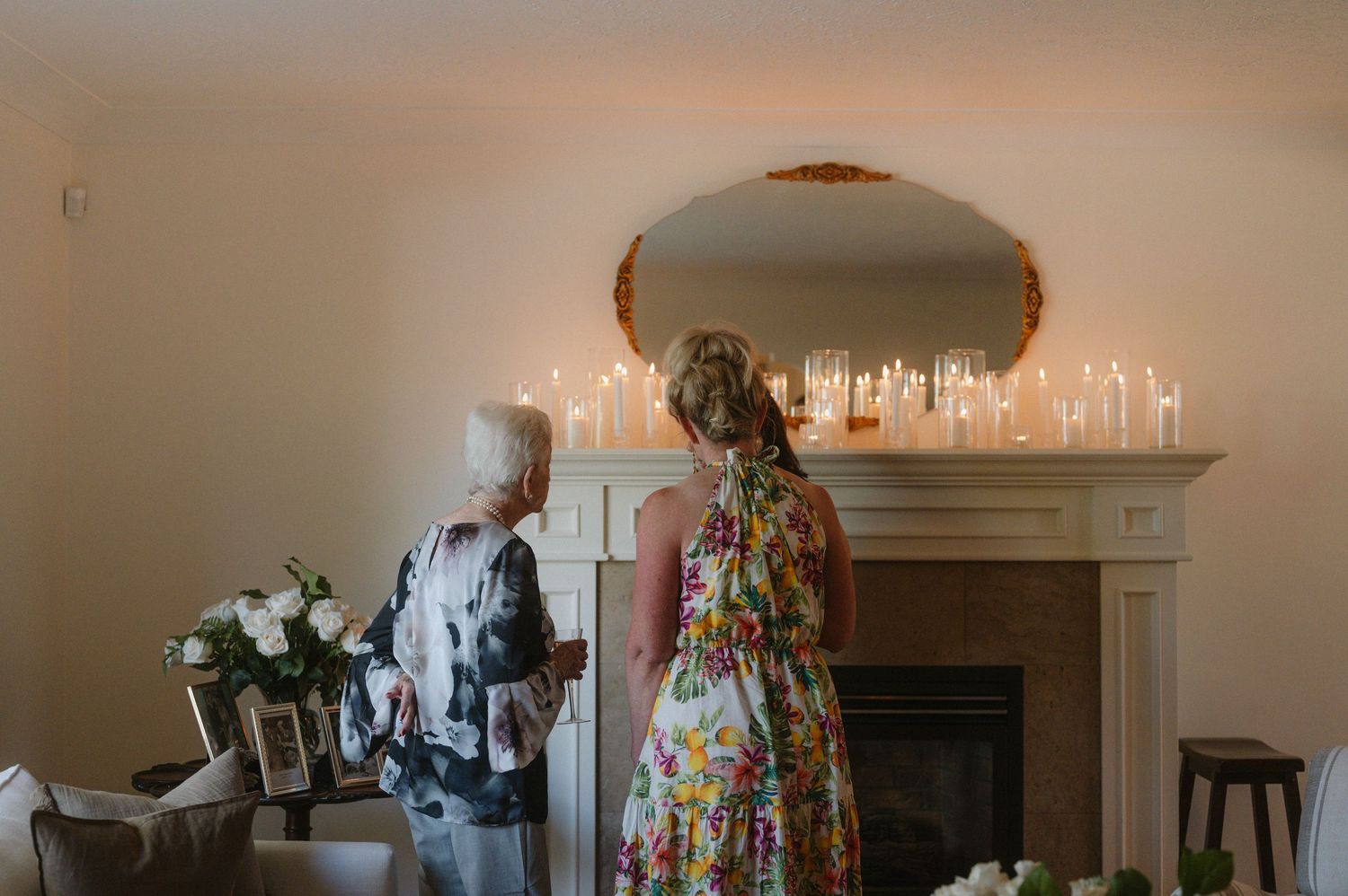 Two people stand in front of a fireplace mantel decorated with glowing candles and white floral arrangements.