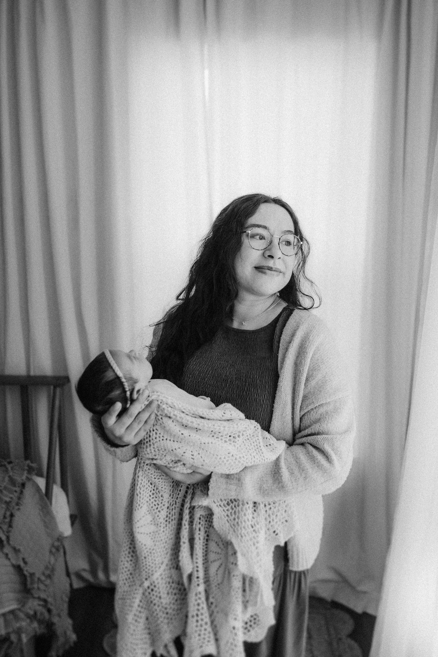 A person wearing glasses and a cardigan holds a knitted blanket while standing against white curtains in a monochrome photo.