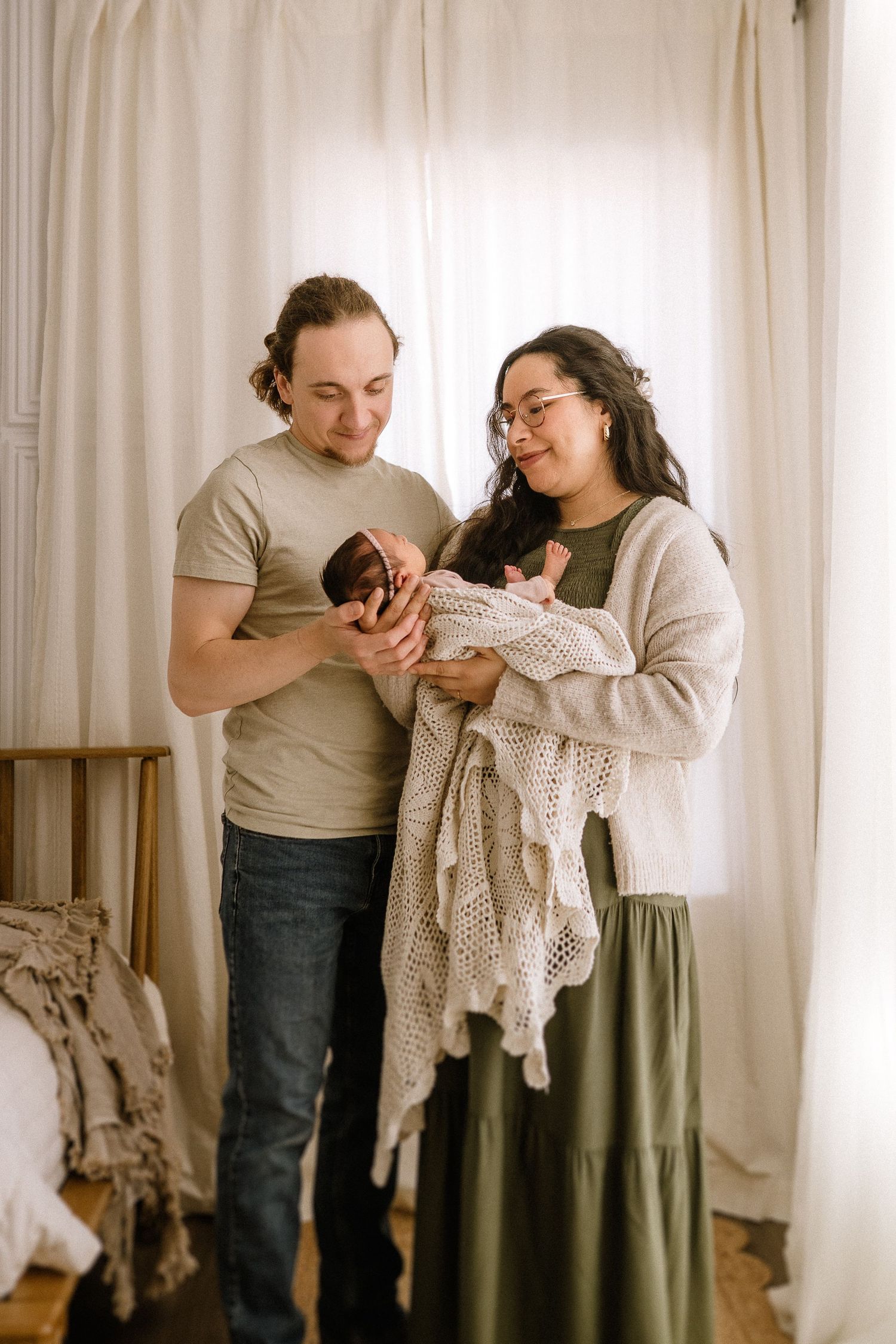 Two people share a tender moment looking down while holding a knit blanket in a softly lit room by white curtains.