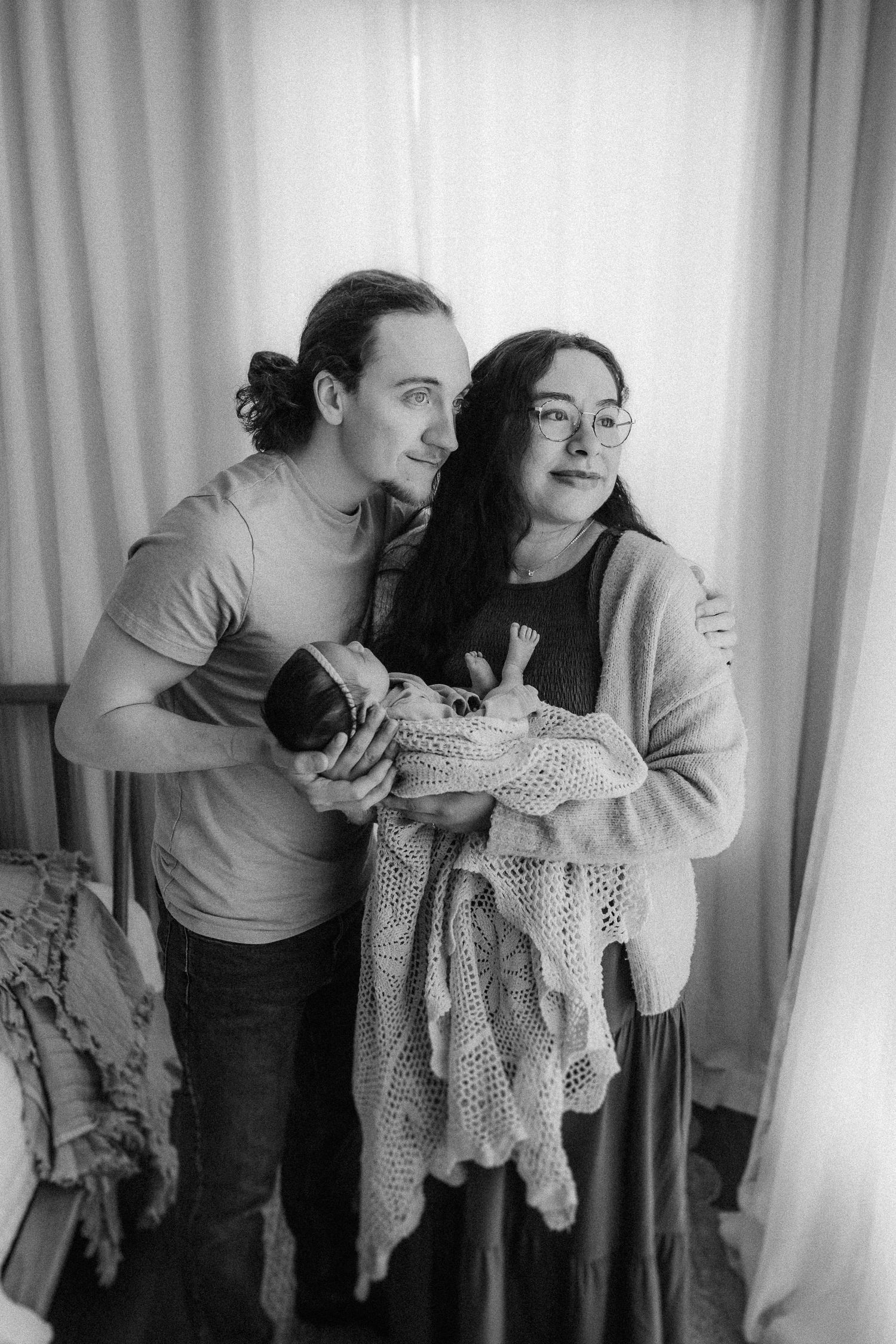 Black and white photo of a couple cradling a newborn baby wrapped in a crocheted blanket near a curtained window.