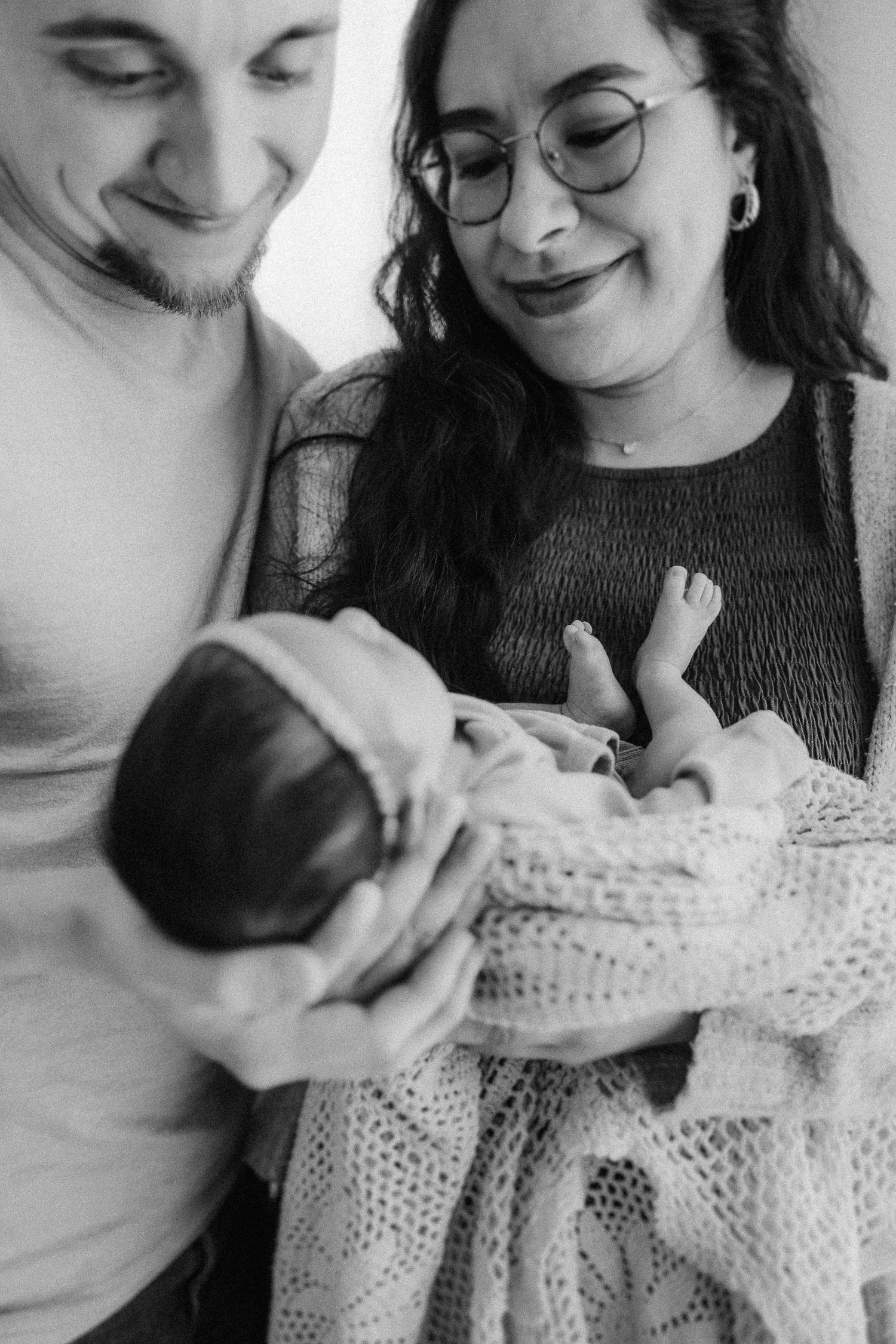 Black and white photo of a couple lovingly gazing down at their newborn baby wrapped in a cozy knit blanket.