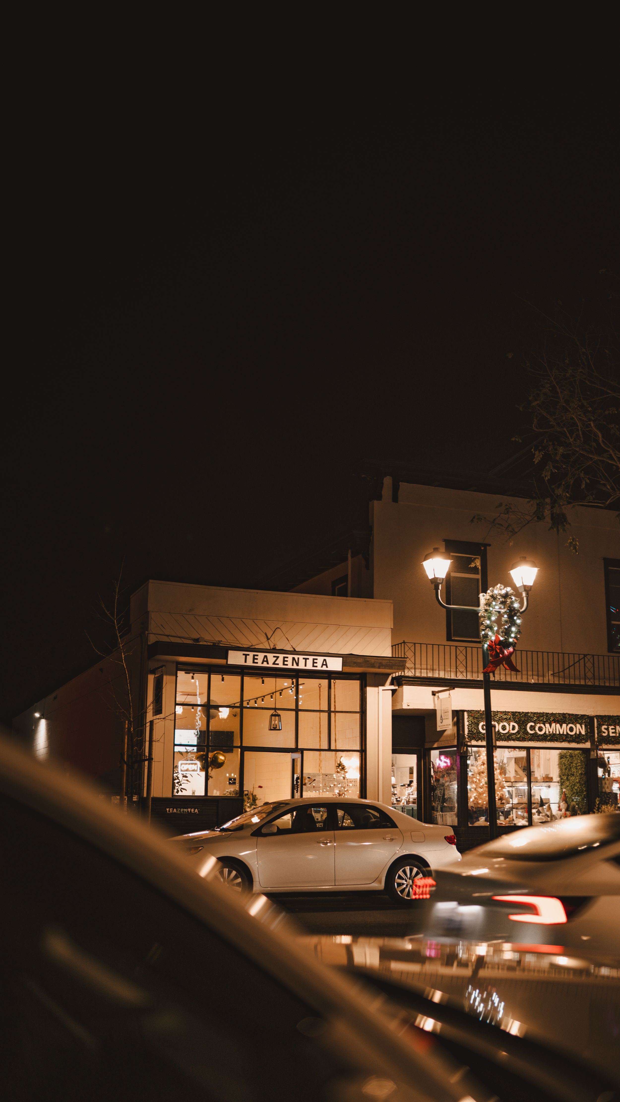 Warm illuminated storefront of a business with a parked car at night and vintage street lamp creating a cozy atmosphere.