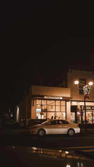 Night view of a small boutique storefront with glowing street lamp and silver sedan parked outside in moody lighting.