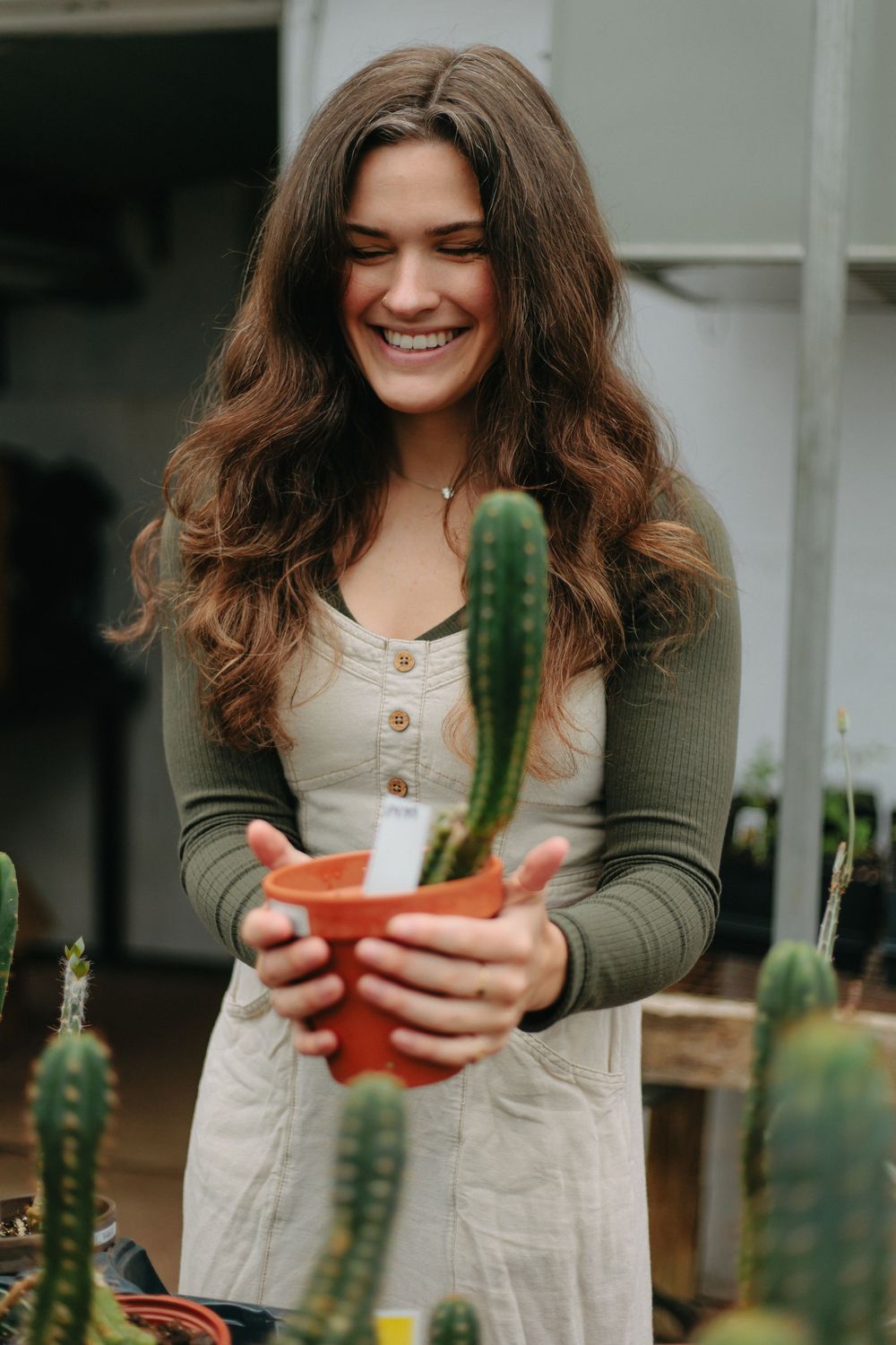 A person in a green cardigan holds a small potted cactus while standing among other succulent plants.