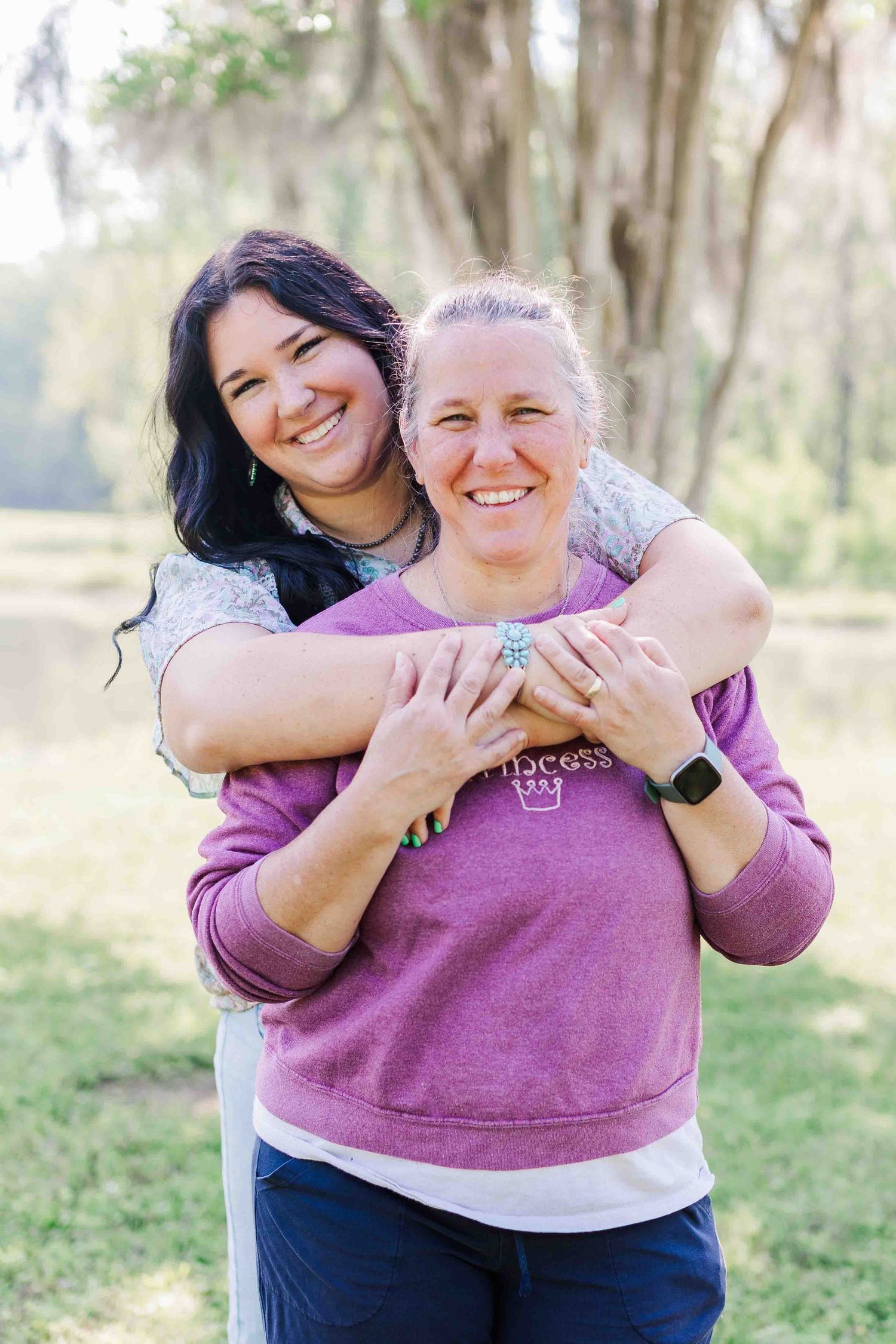 Two friends share a joyful embrace while standing outdoors in a sunlit wooded area.
