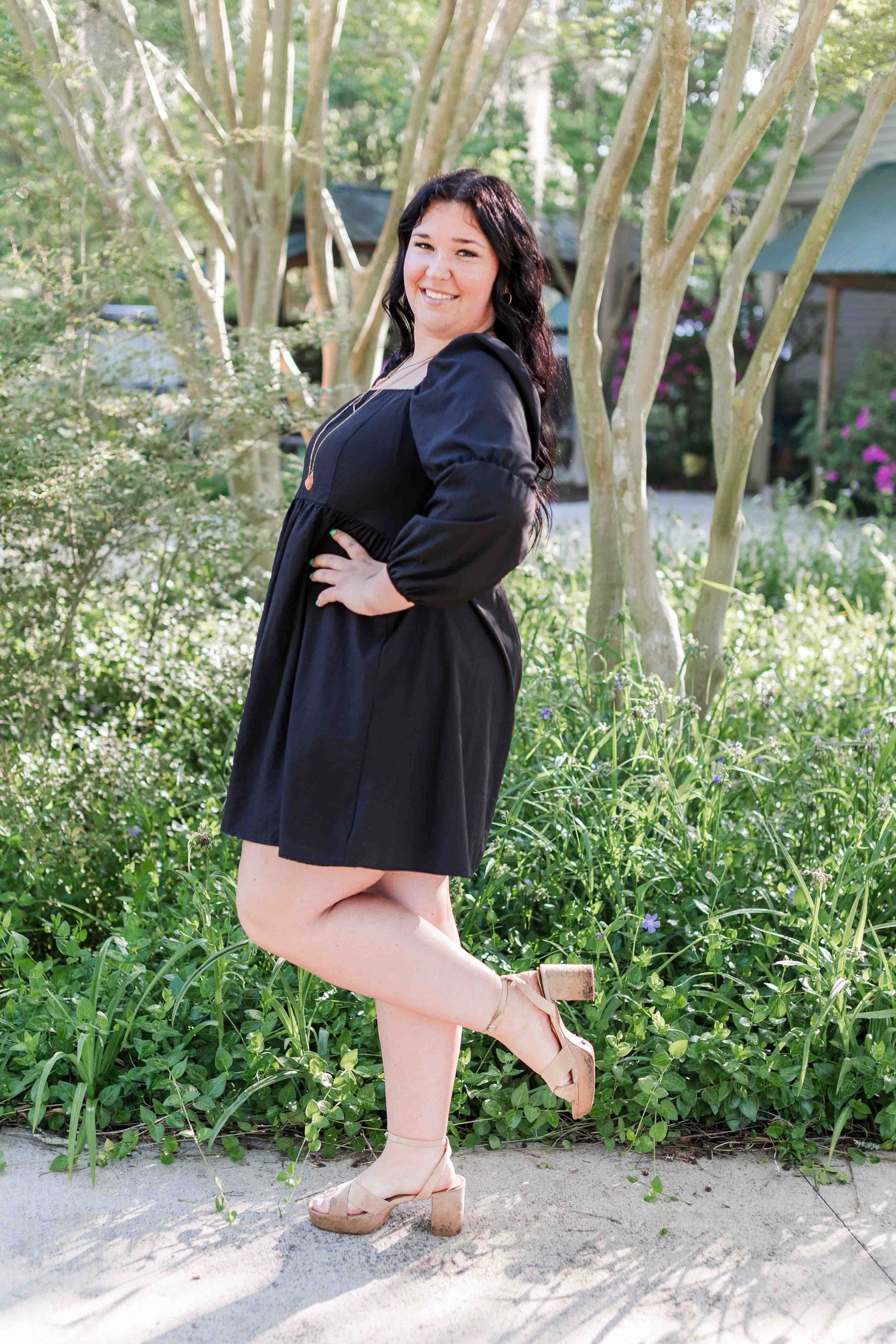 Person in a black dress posing outdoors against greenery with decorative trees and landscaping in the background.