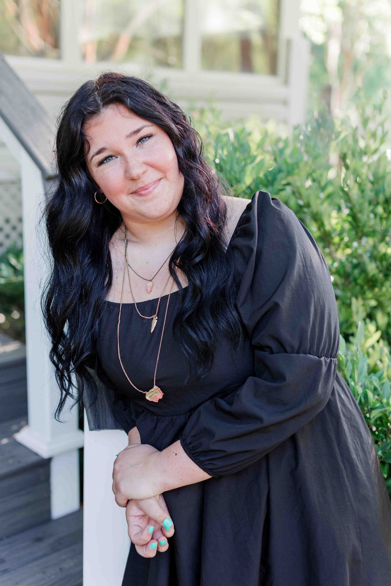 A woman in a long-sleeved black dress stands outdoors near green foliage and a white railing.
