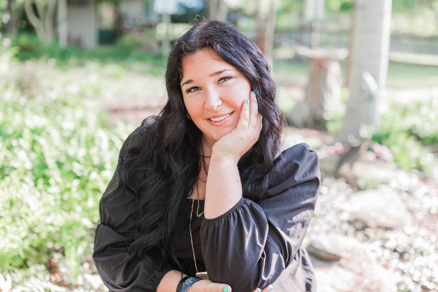 Long dark haired person in black blouse smiling while resting chin on hand outdoors in natural setting.