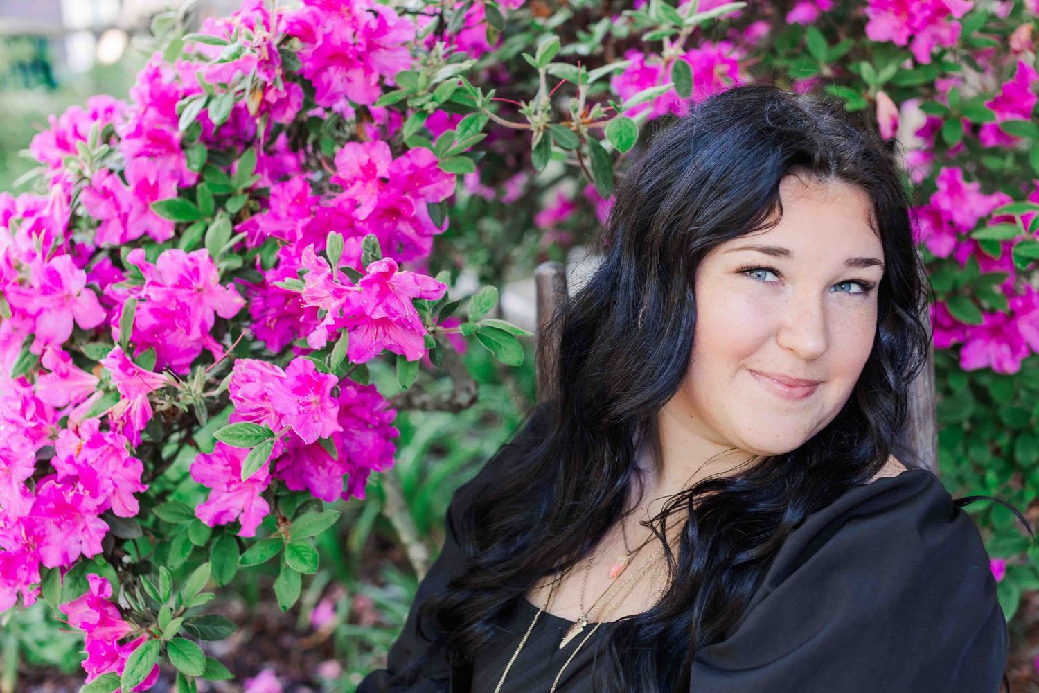 A dark-haired person in a black top poses near vibrant pink bougainvillea flowers in an outdoor setting.