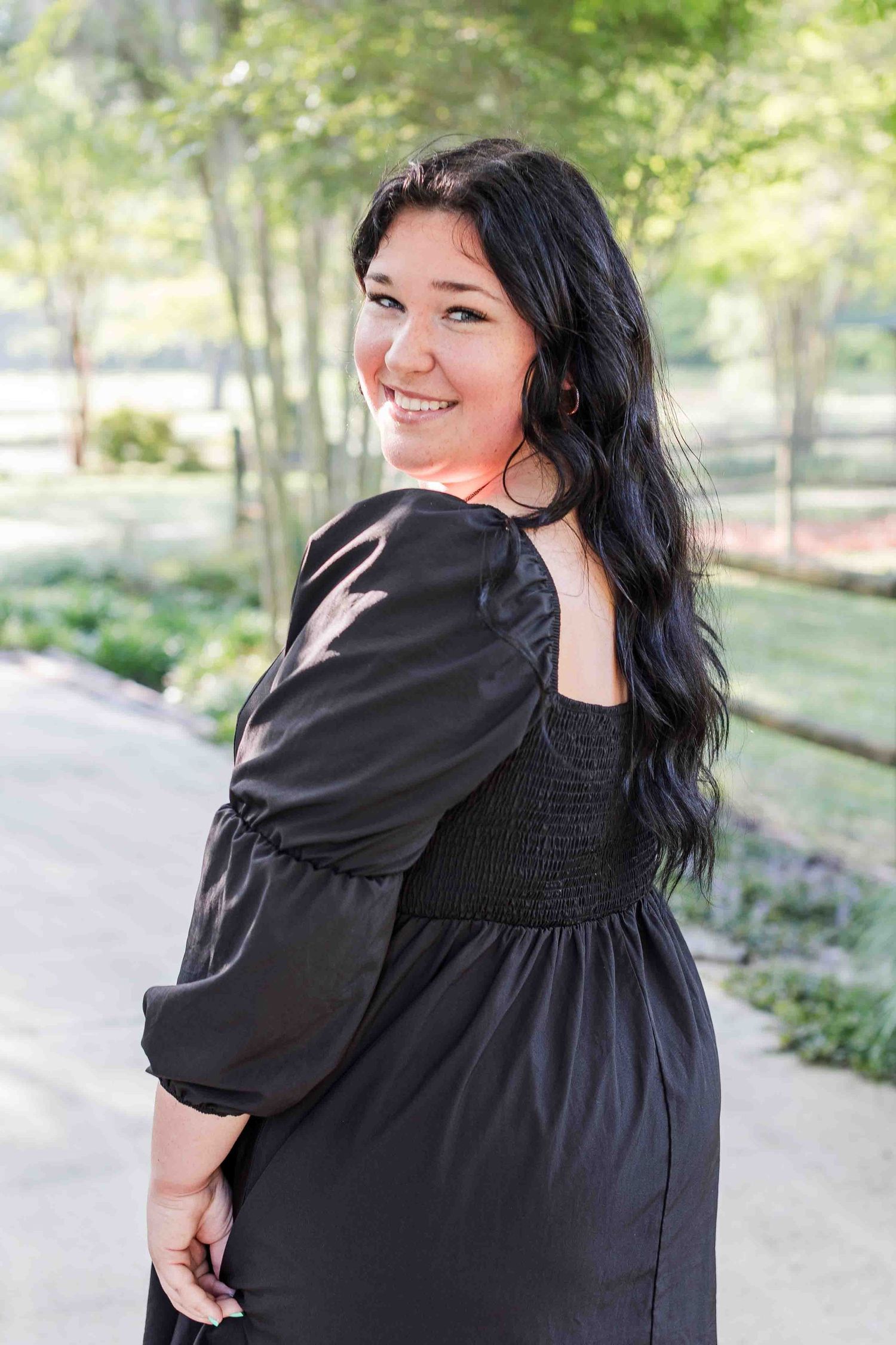 A woman in a black dress smiles while looking over her shoulder in a park setting on a sunny day.