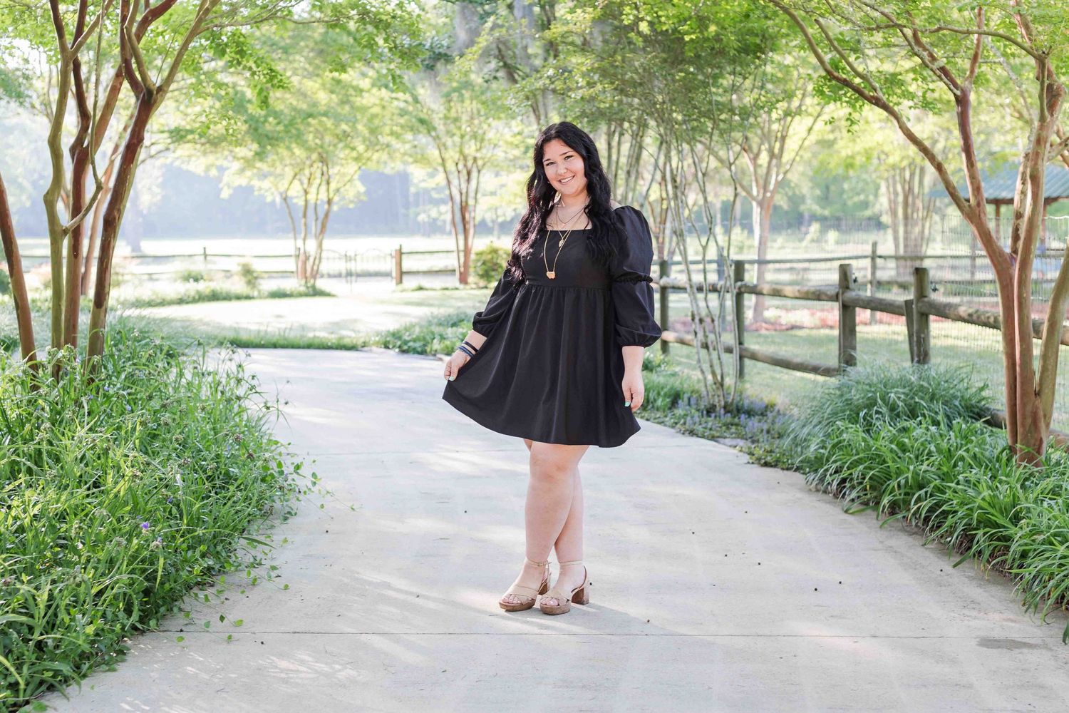 Person in a black dress poses on a tree-lined pathway in a landscaped garden setting with green foliage.