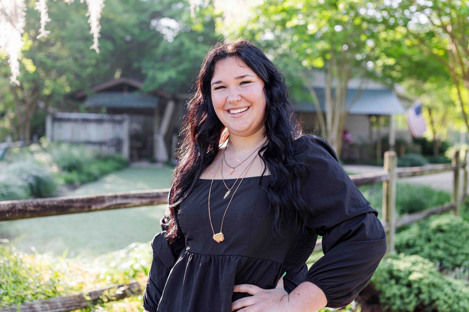 Woman in black peasant dress with long dark hair poses outdoors in a garden setting with natural lighting.