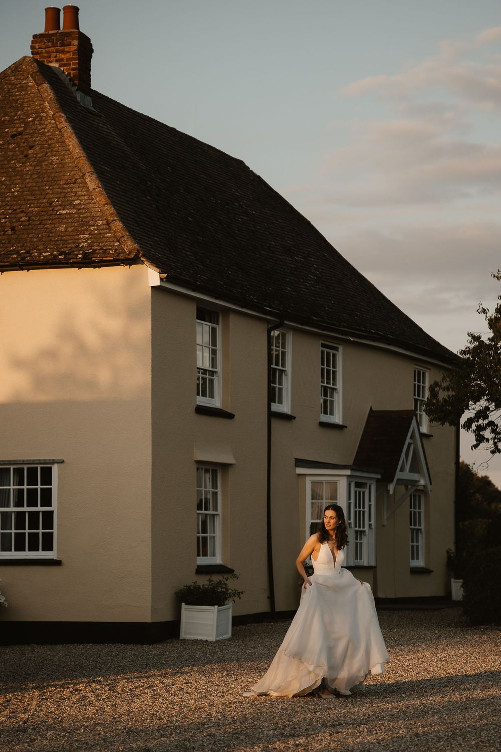 Evening sunset light illuminates a series of white cottage buildings with sash windows and brick chimneys along a gravel path.
