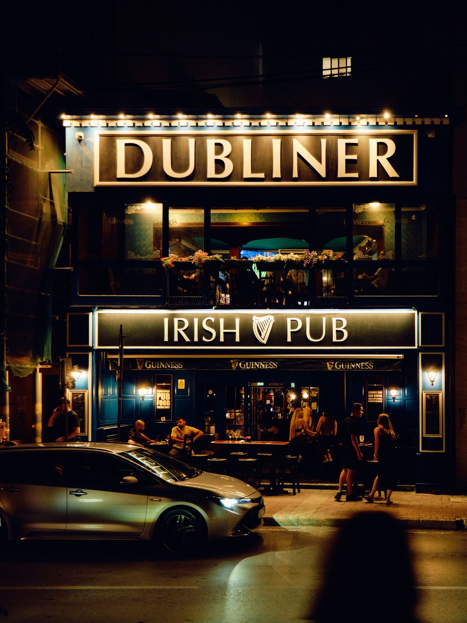Illuminated storefront of a Dubliner Irish Pub at night with a parked car and pedestrian silhouette visible.