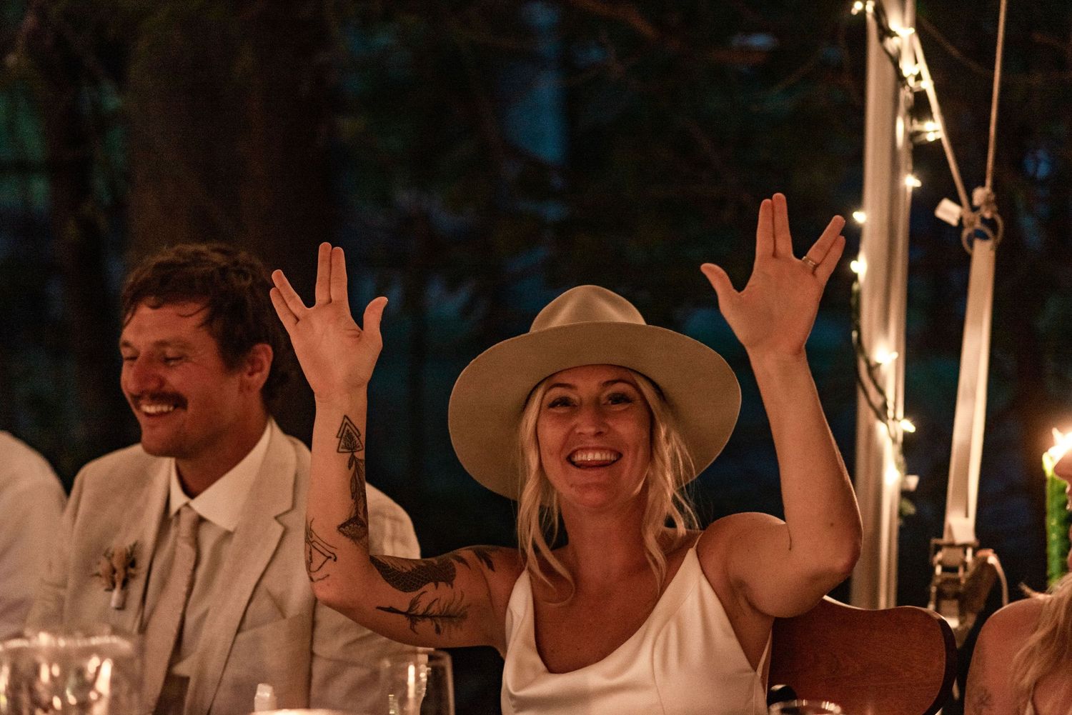 Wedding guests raise their hands in celebration during an outdoor evening reception with string lights overhead.