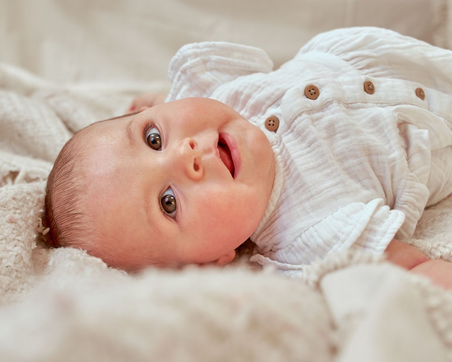 Baby in white outfit lying on soft fabric.