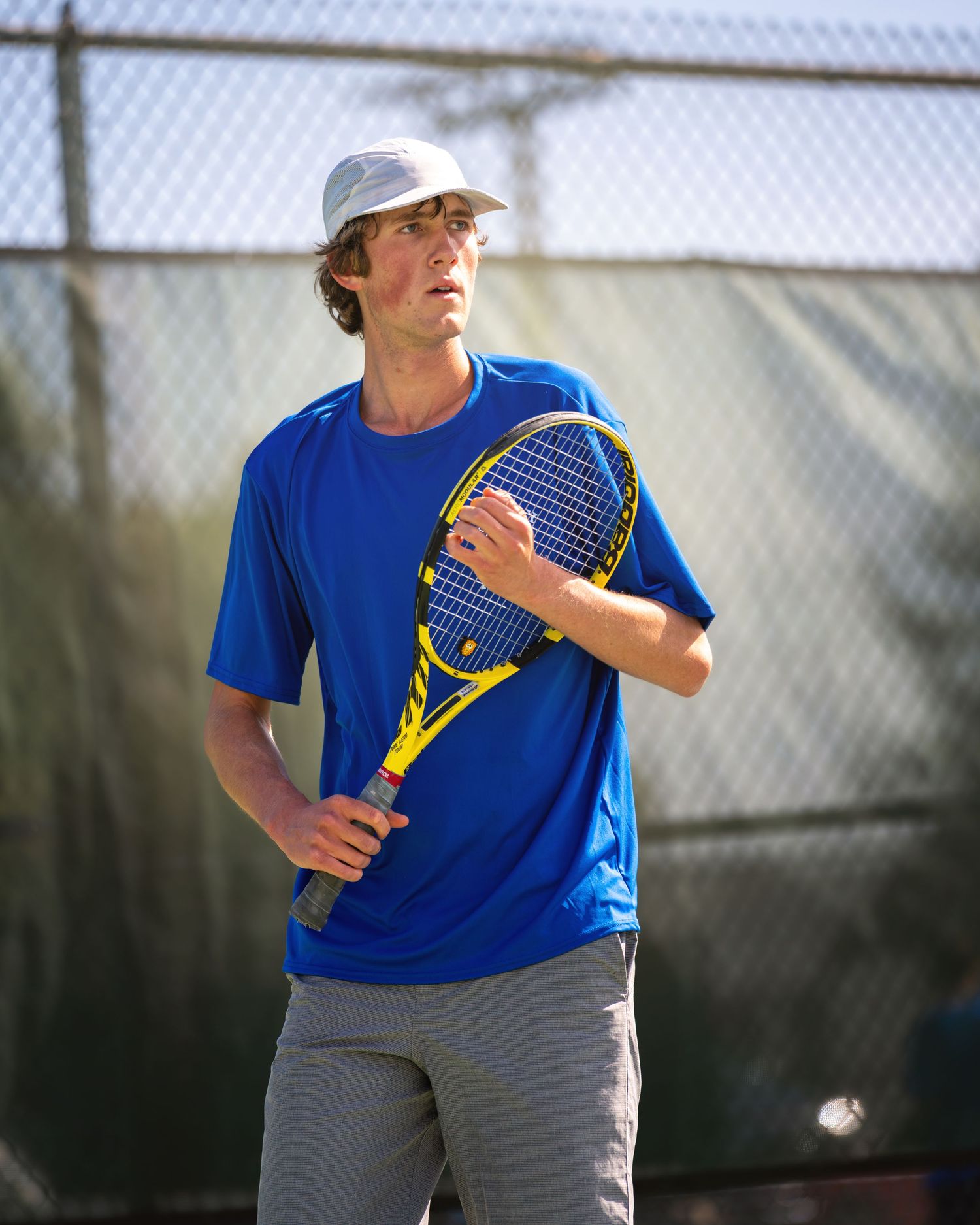 Tennis player in bright blue shirt and baseball cap holds yellow racquet while standing on outdoor court.