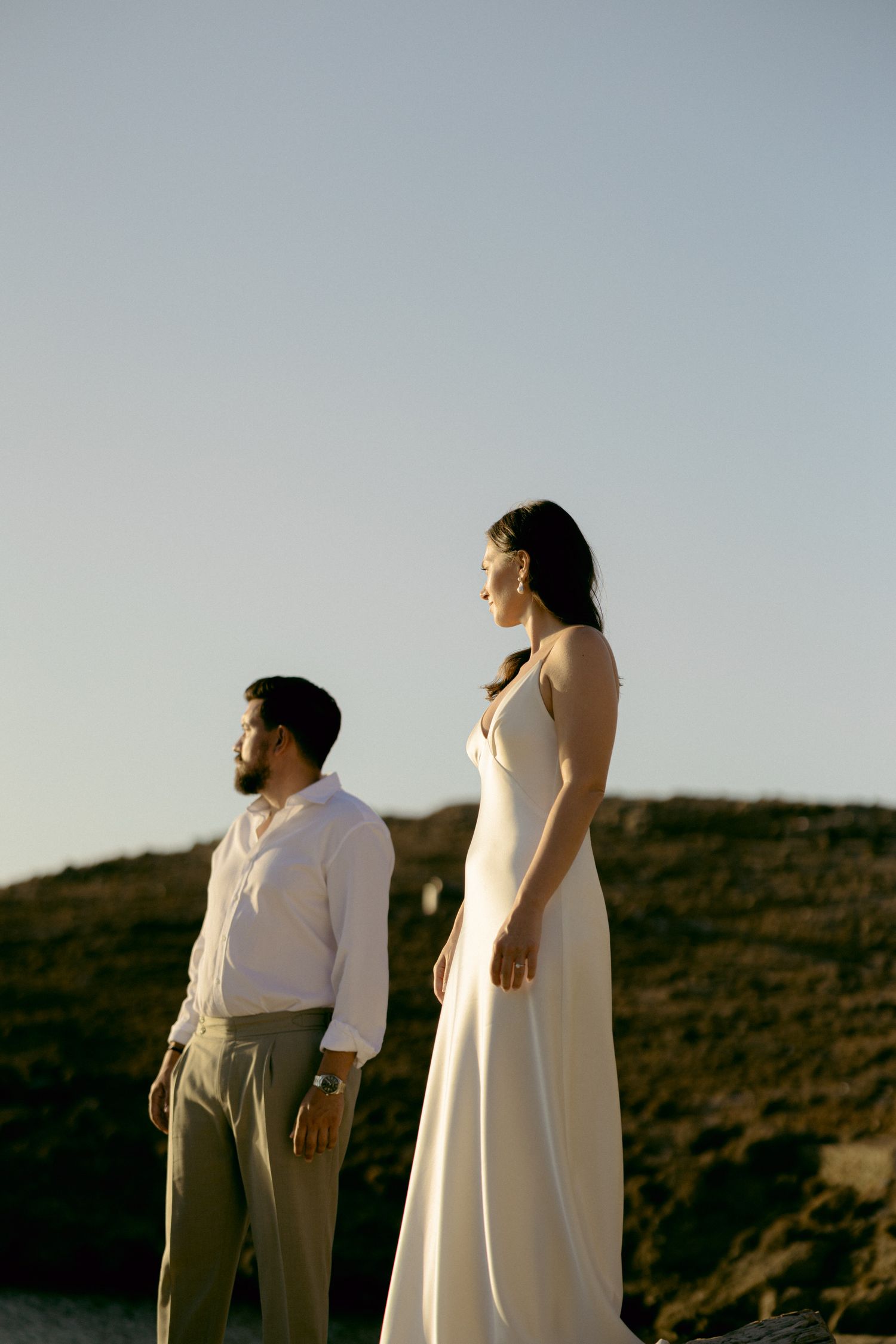 A couple stands atop a hill at golden hour in elegant white wedding attire in Kythnos.